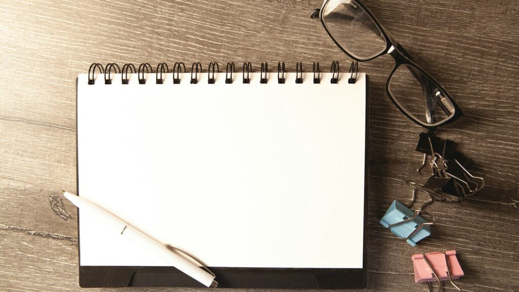 A blank notepad on a wooden desk beside a pen, glasses and paper clips
