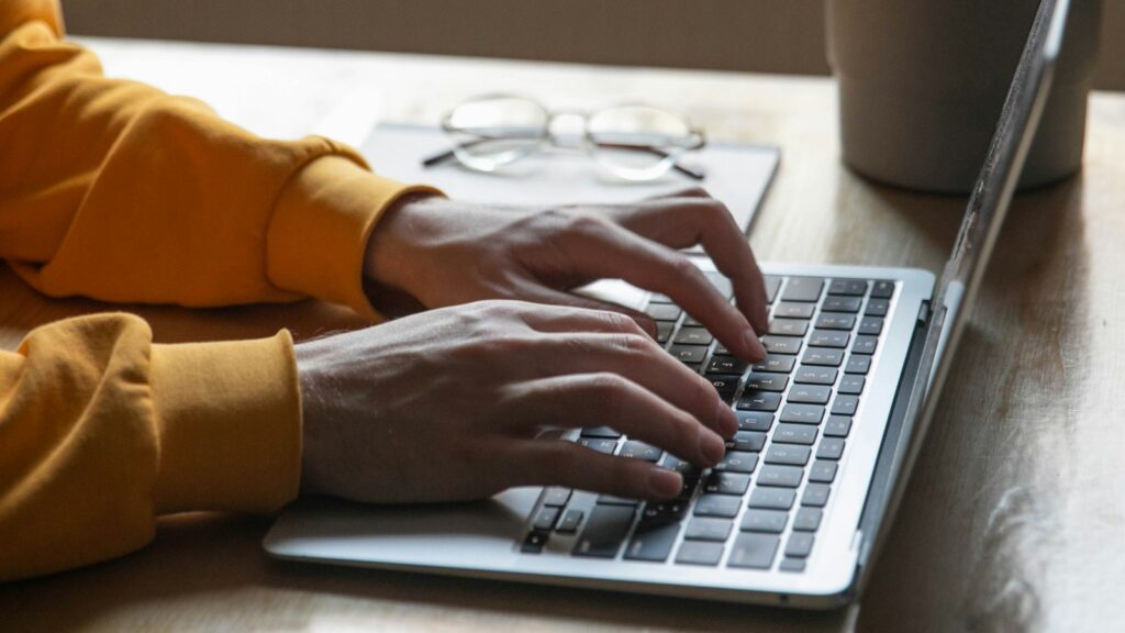 A person in a mustard sweater typing on a laptop beside a small plant